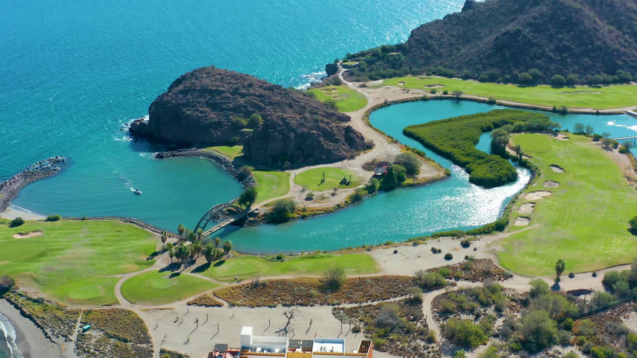 High oblique aerial over Loreto, Baja California Sur showing the coastal lagoon, open ocean, rocky headland, green field with sand traps, shore road, and no visible people.