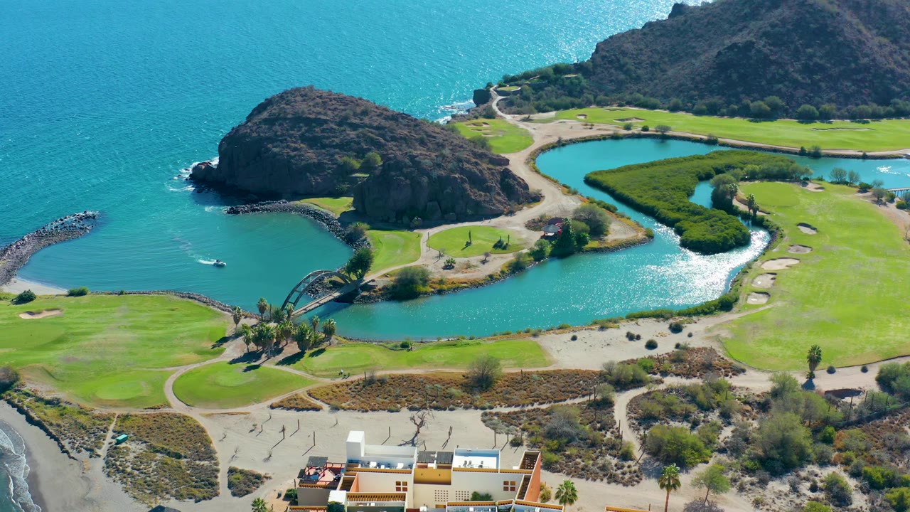 Aerial frame over Loreto, Baja California Sur with the golf lake centered, fairways around it, a rocky hill at left, ocean behind, and no visible people.