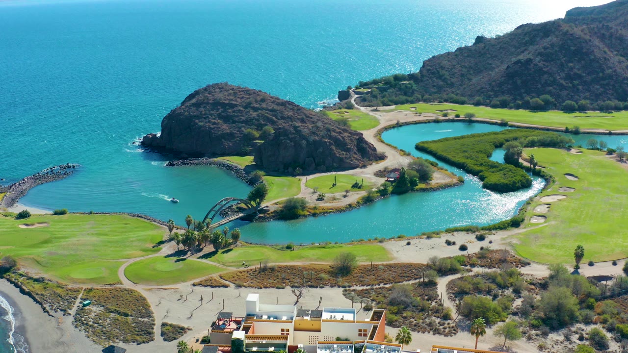 Opening aerial frame over Loreto, Baja California Sur shows a turquoise lagoon beside the ocean, green grounds, rocky hills, beach surf, and low buildings at the bottom edge, with no visible people.