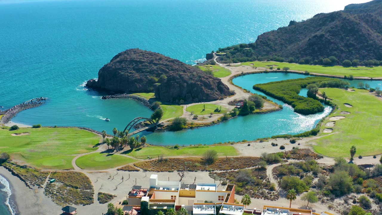 At 0:02, the frame shows the rocky headland above the lagoon, bright green shoreline grounds, blue ocean water, and a strip of buildings along the bottom, with no visible people.