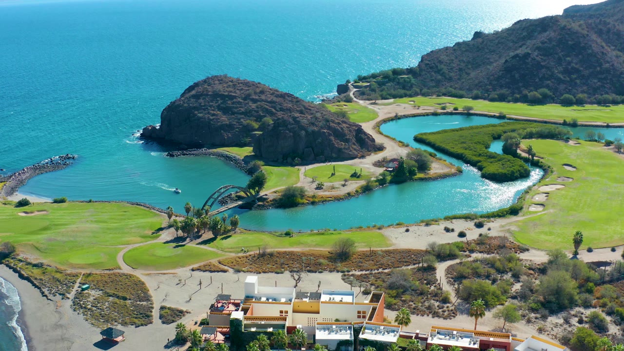 At 0:03, the Loreto coastline remains framed from above with the lagoon centered, white waves curling on the right beach, ocean behind the rocky hill, and no visible people.