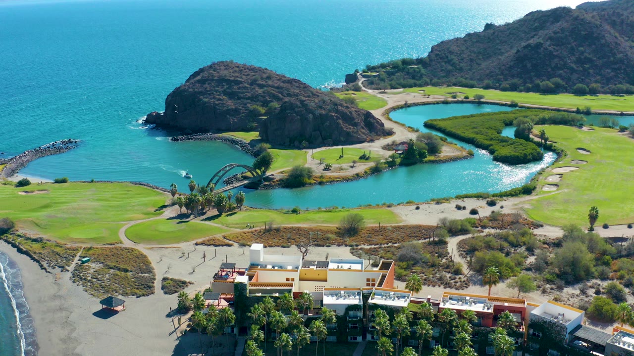 Wide aerial frame over Loreto, Baja California Sur showing ocean water, a rocky coastal outcrop, green golf fairways, a curved lagoon, shore buildings, and no visible people.