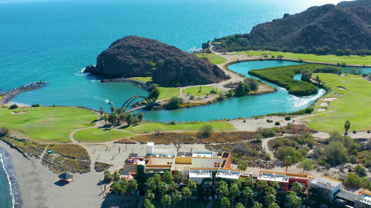 Drone view of lagoon water curving around golf greens beside a rocky headland and ocean.