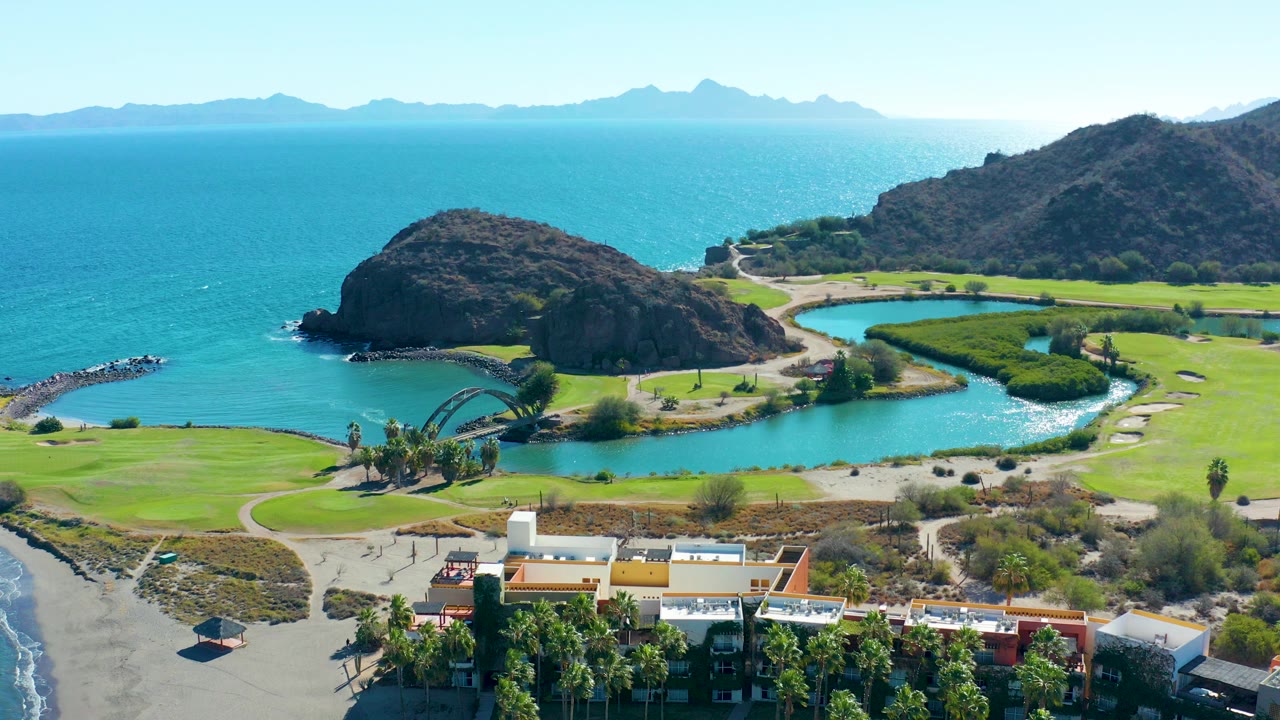 Aerial frame over Loreto, Baja California Sur showing turquoise ocean, a curved lagoon, green coastal grounds, sandy beach, rocky hills, distant mountains, foreground buildings, and no visible people.