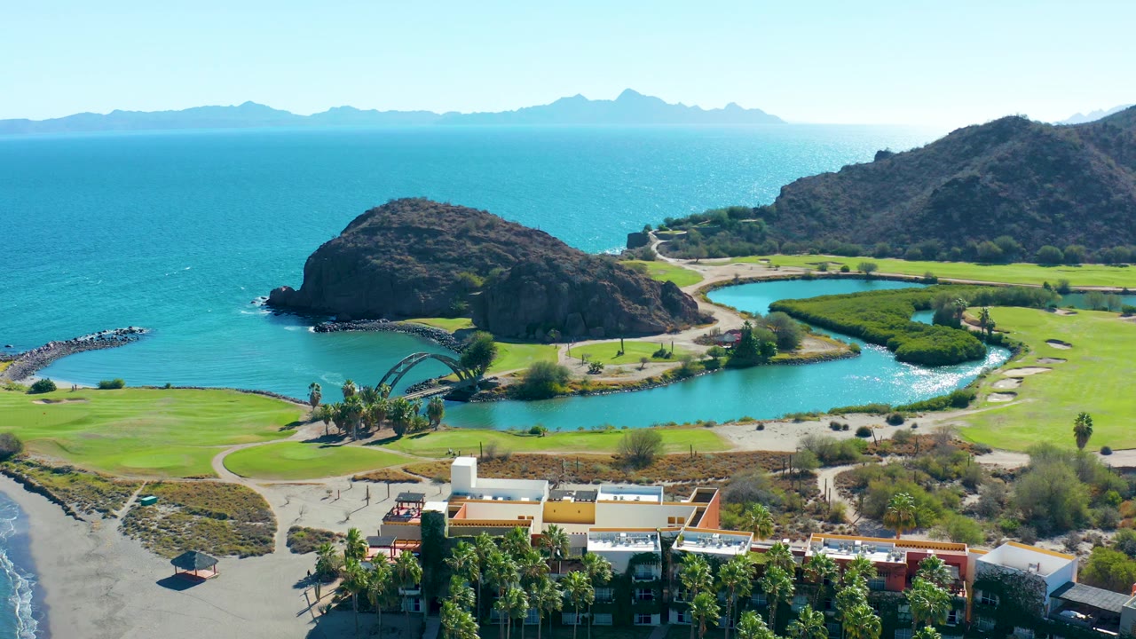 A bright aerial frame from Loreto, Baja California Sur showing the lagoon curving beside green grass, ocean water beyond the beach, dark rocky hills, distant mountain silhouettes, and no visible people.