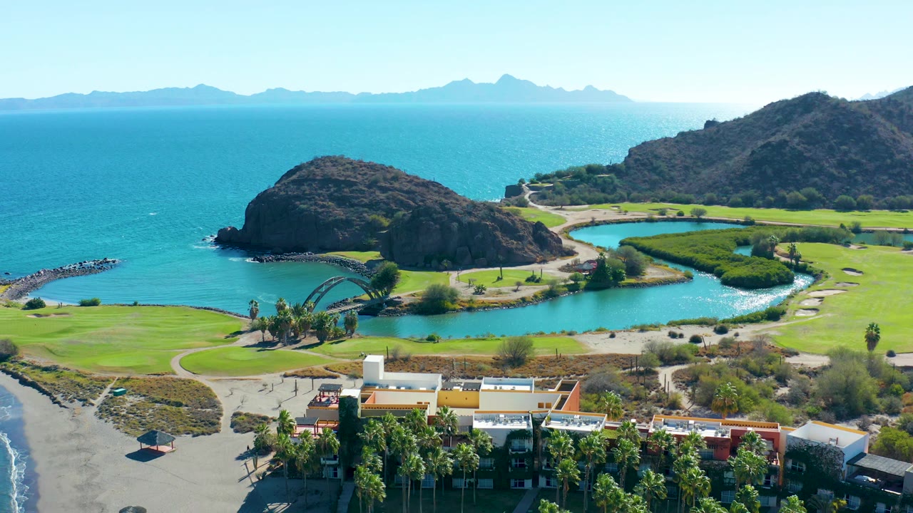 Aerial coastal landscape with ocean, rounded rock hill, lagoon, beach, and foreground rooftops.