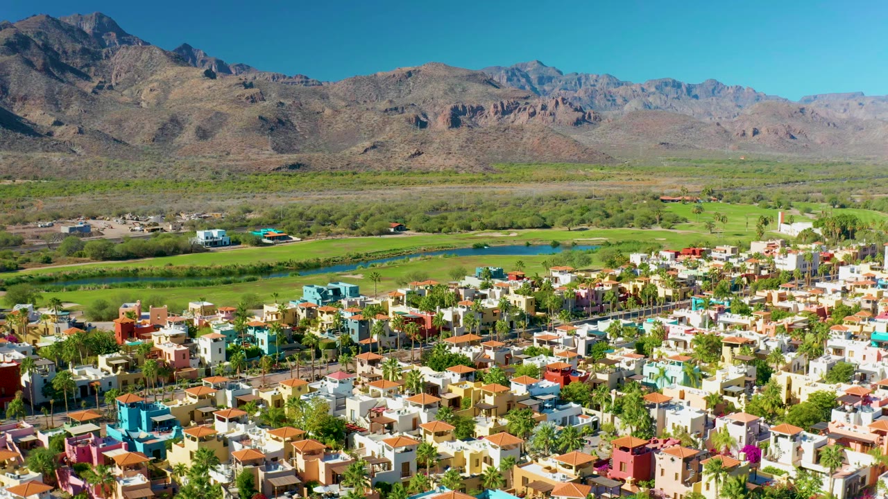 Aerial opening frame over colorful residential homes in Loreto, Baja California Sur, with a water channel, green fields, brown mountains, clear blue sky, and no visible people.
