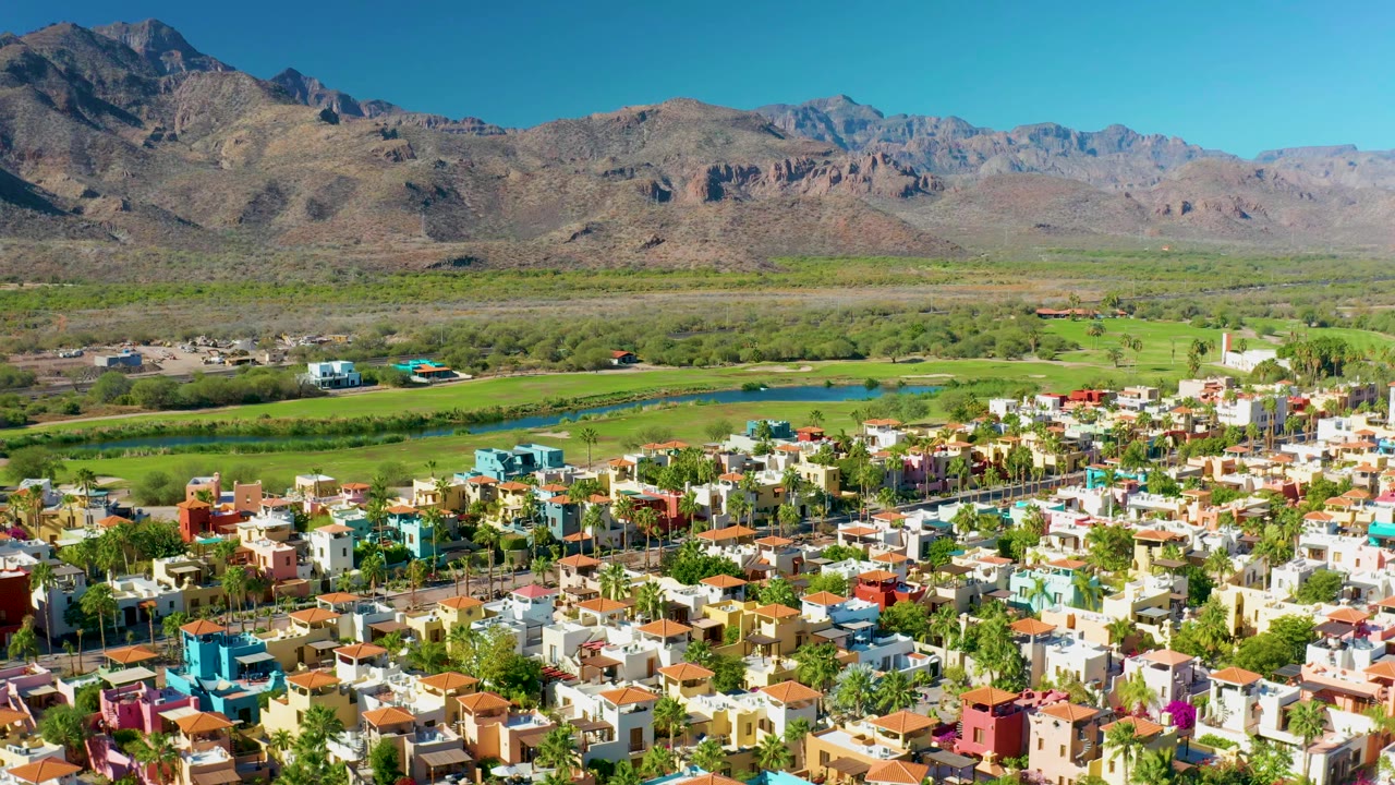 Elevated frame over Loreto residential rooftops, with white and pastel houses in the foreground, palms, a blue-green river channel, fields, mountains, and no visible people.