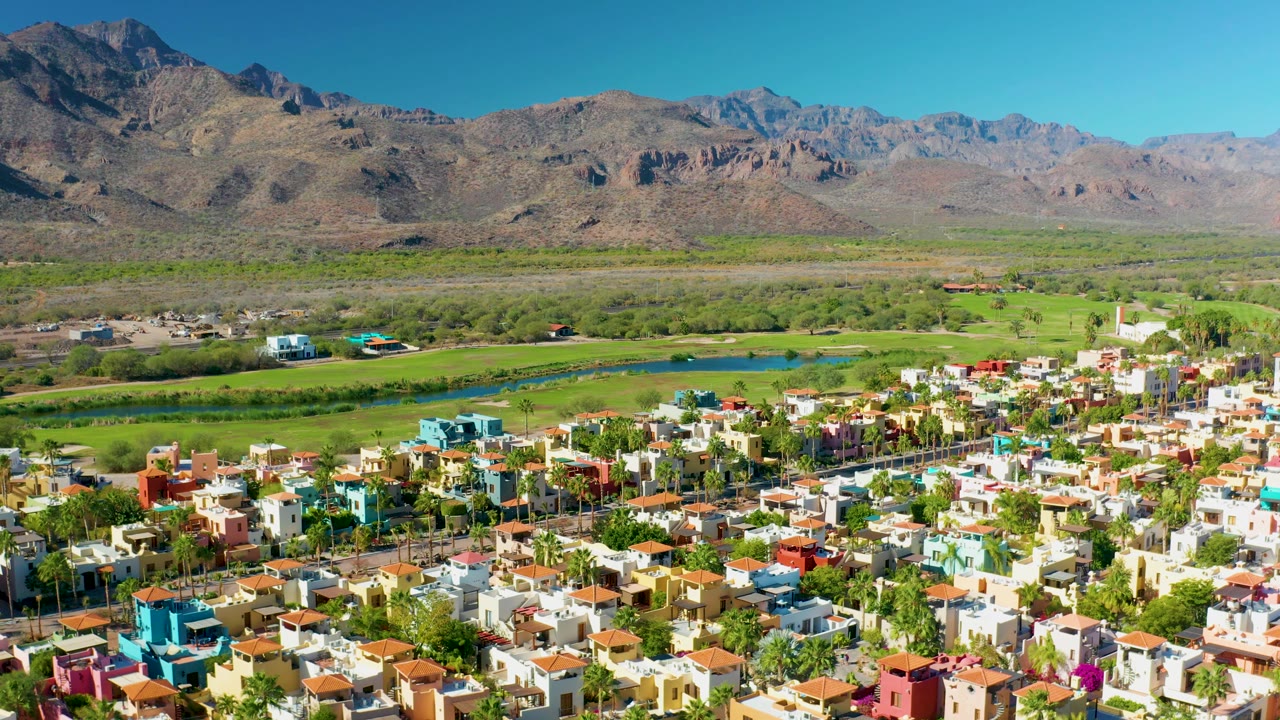 Aerial view across a colorful Loreto neighborhood, showing dense low-rise homes, tree-lined streets, a narrow waterway, green fields, distant mountain ridges, and no visible people.