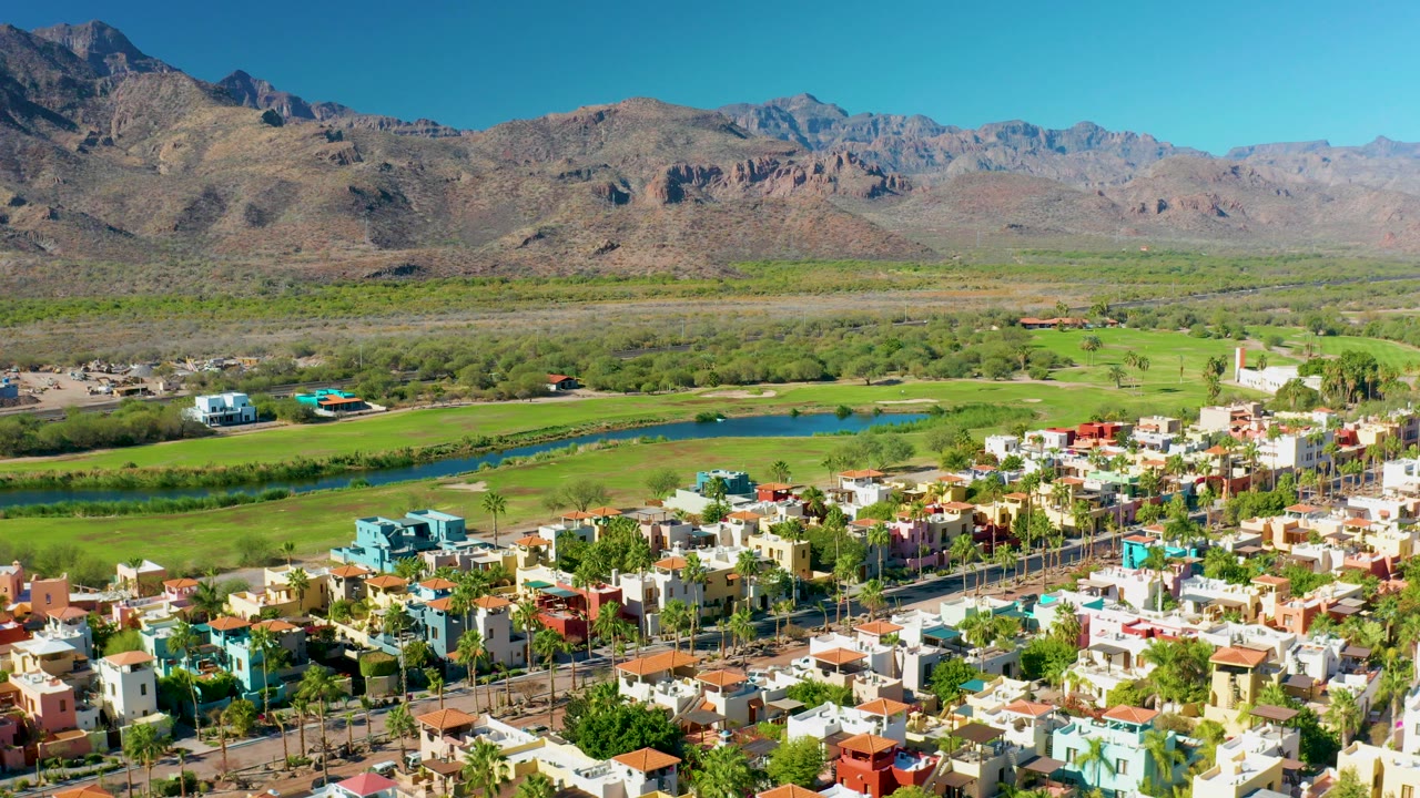 Opening aerial frame over Loreto, Baja California Sur with colorful homes in the foreground, a narrow river and green fields beyond, dry mountains in back, and no visible people.