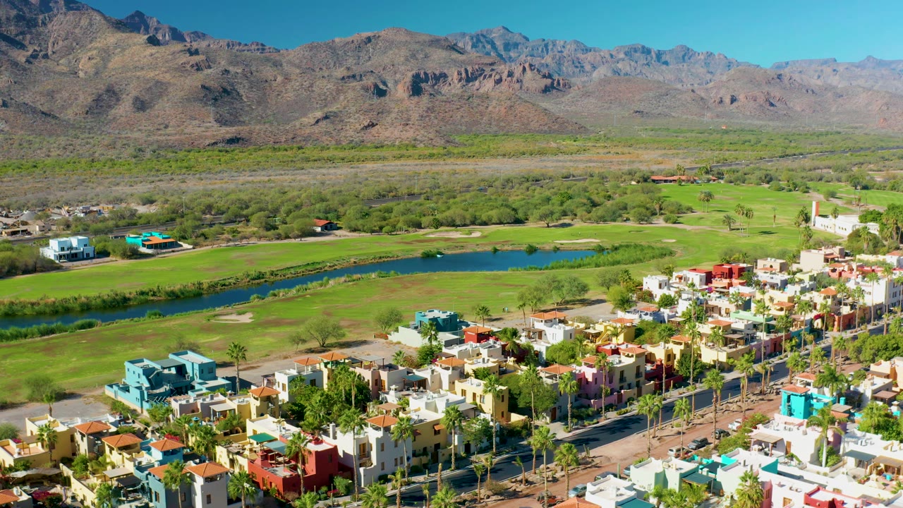A slightly later aerial view in Loreto, Baja California Sur keeps the colorful housing blocks low in frame, with fairways, a blue water strip, desert brush, mountains, and no visible people.