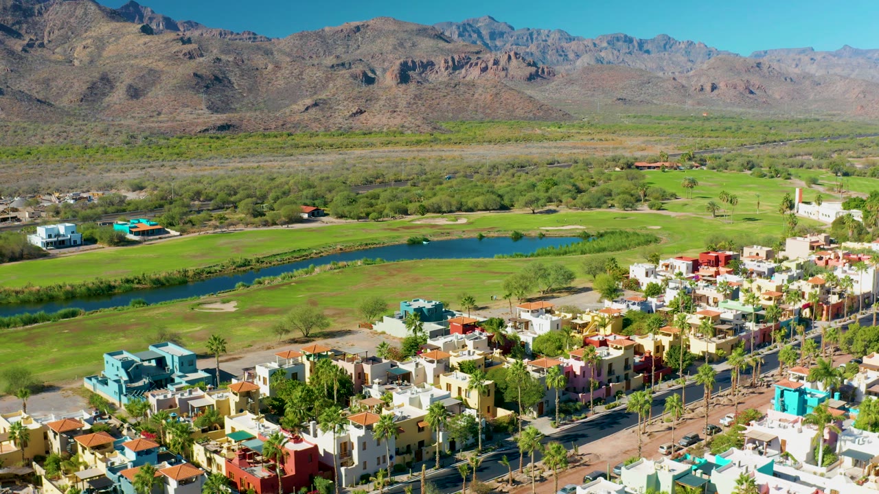 The aerial composition over Loreto, Baja California Sur shows a palm-lined street and clustered colorful homes at the lower right, green golf course terrain across the middle, mountains beyond, and no visible people.
