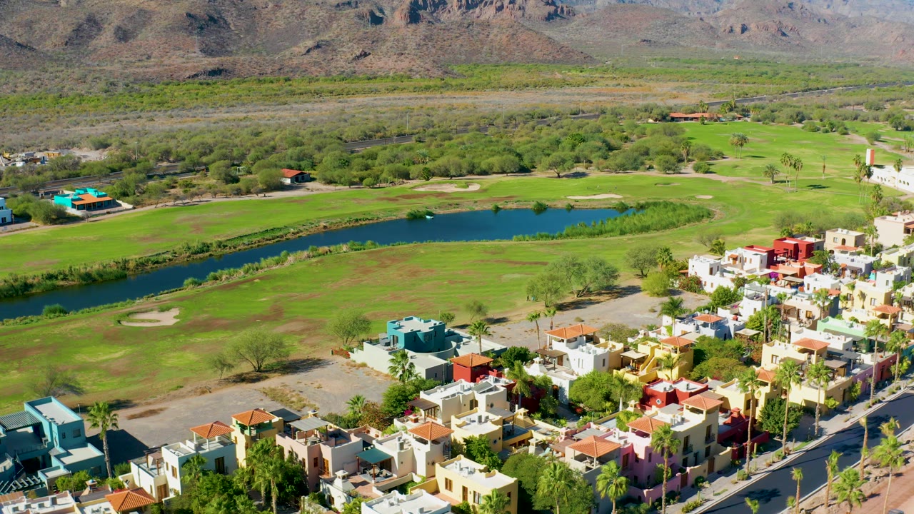 Aerial view over Loreto homes and roads with the golf course pond running across the middle of the frame, green fairways behind it, mountains on the horizon, and no visible people.