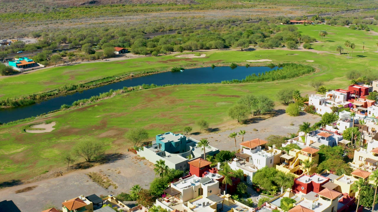 High aerial frame over a Loreto, Baja California Sur golf course with a blue pond across the middle and homes with red and white roofs in the lower right.