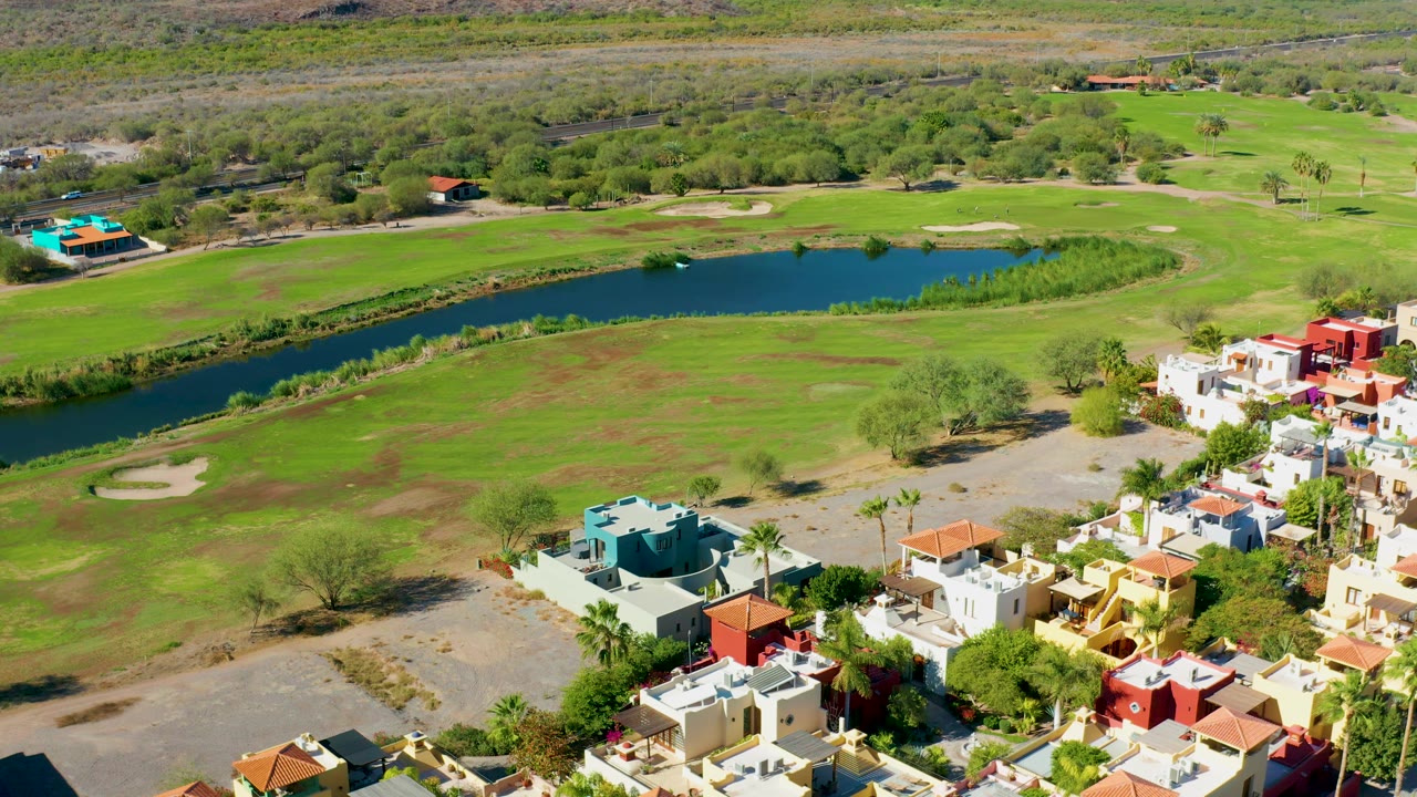 Aerial view across fairway grass, a pond, and houses at frame edge.