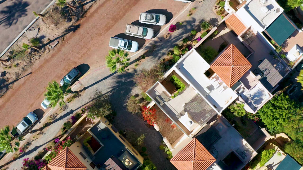 Top-down aerial frame over Loreto, Baja California Sur, showing a reddish residential road, several parked cars, white houses with red tile roofs, palms, garden walls, and no visible people.