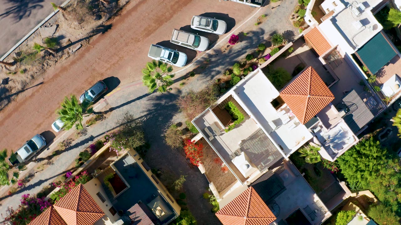 Aerial residential block with a reddish road, parked cars, palms, and house roofs.