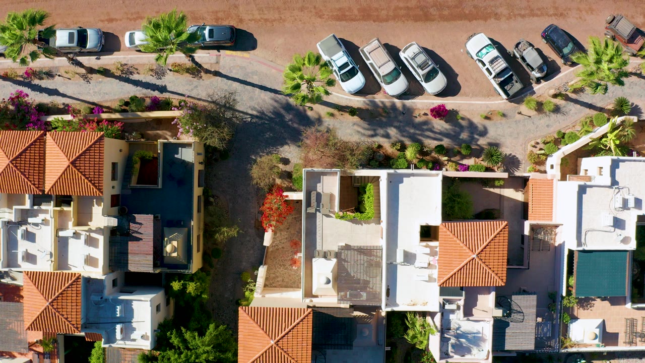 Overhead drone frame in Loreto, Baja California Sur showing white and tan homes, red tile roofs, parked cars, palms, garden greenery, and no visible people.