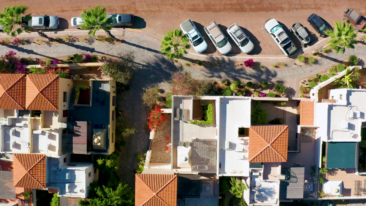 Top-down residential block with white rooftops, red tile awnings, cars, palms, and shadows.