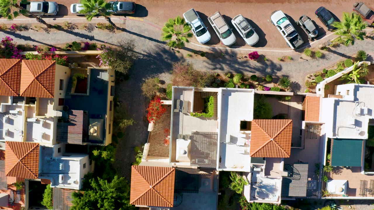 Drone frame over Loreto homes with a central white roof, red clay roof panels, enclosed courtyards, parked cars along the upper road, and no visible people.