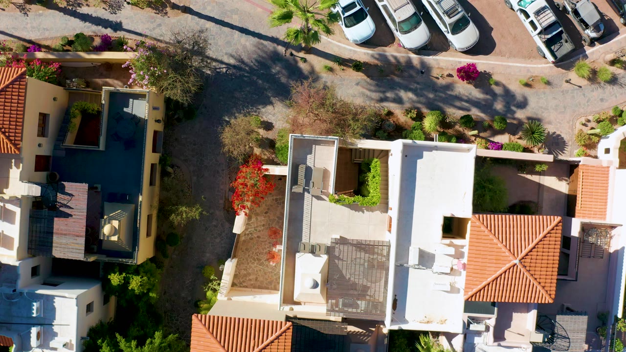 Straight-down aerial view of residences, parked vehicles, bright roofs, and street landscaping.