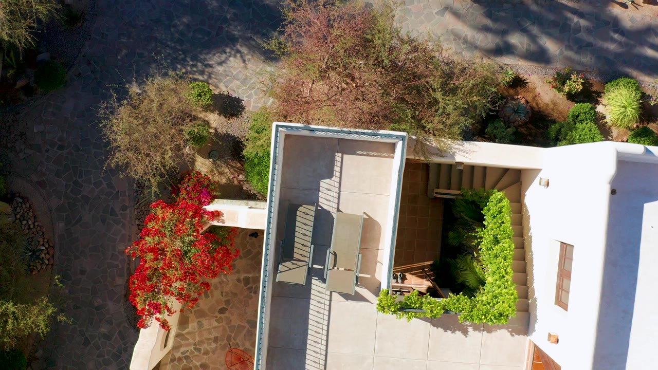 Top-down view in Loreto, Baja California Sur showing a white house patio, chairs, red flowering bush, green hedge, stone path, and no visible people.