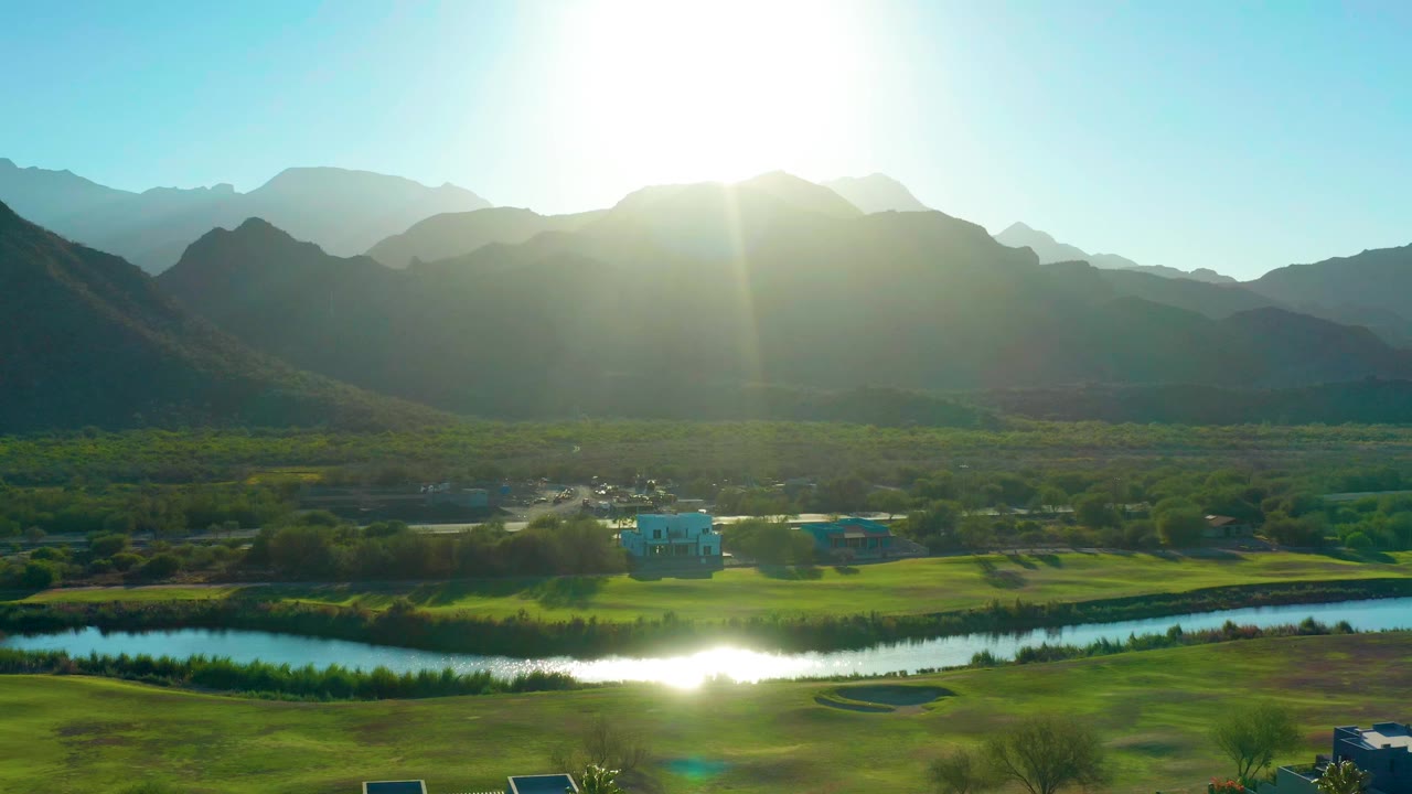Wide aerial view in Loreto, Baja California Sur, with a bright river bend, green fields, a pale blue building, mountain ridges, and no visible people.