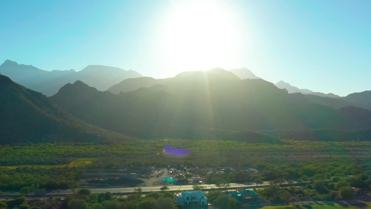 Second frame of Loreto, Baja California Sur with the sun blown out above the ridgeline, blue sky overhead, green vegetation across the valley floor, a road line below, and no visible people.