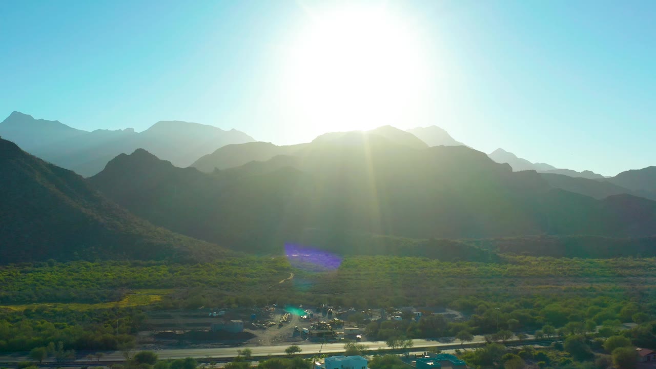 Third frame of Loreto, Baja California Sur showing layered mountain ridges in haze, a green valley with visible roads and scattered structures near the lower edge, bright sun flare, and no visible people.