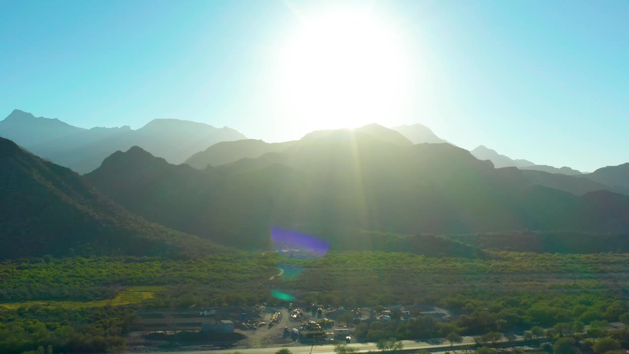 Mountain valley aerial with sun above ridgeline and settlement at bottom.