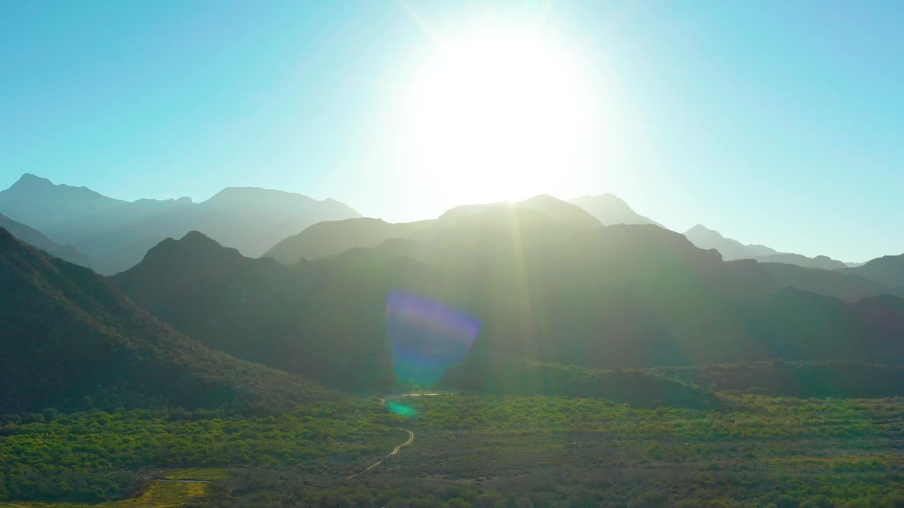 Bright sun hangs above layered mountain ridges in Loreto, Baja California Sur, with a green valley floor, winding dirt road, lens flare, and no visible people.