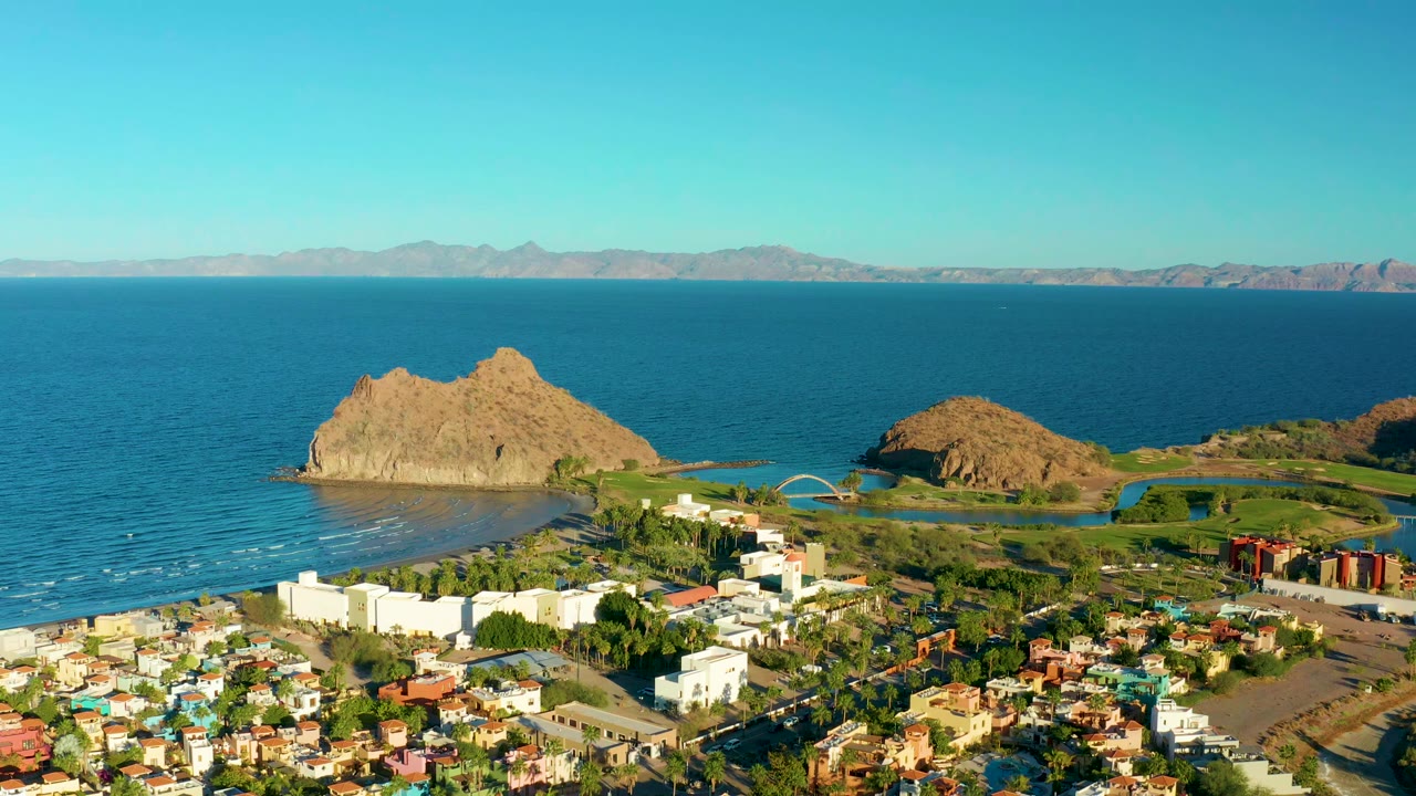 Aerial view in Loreto, Baja California Sur with the rocky hill centered higher in frame, white buildings along the lower shoreline, blue ocean behind it, and no visible people.