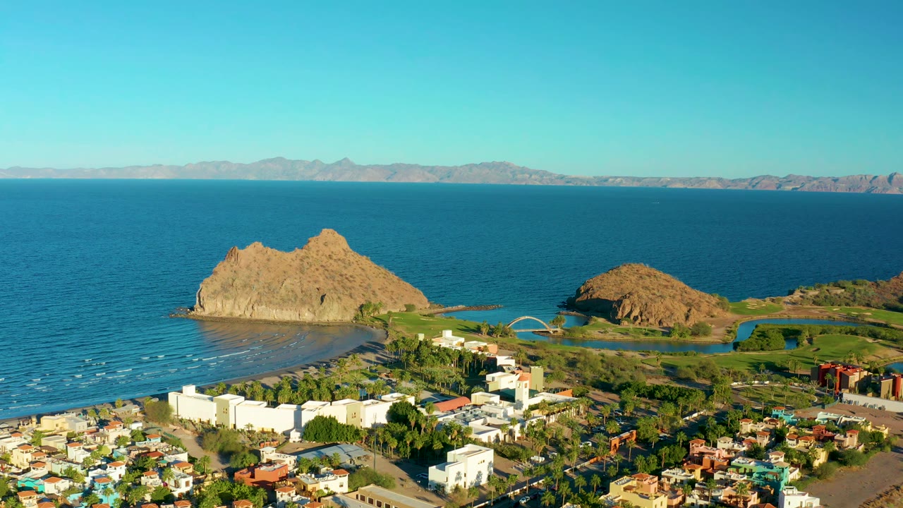 Aerial view over Loreto with the larger rocky outcrop left of center, a smaller outcrop to the right, calm ocean behind them, town rooftops below, and no visible people.