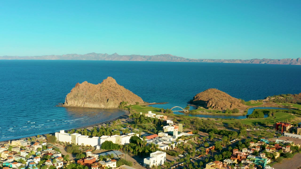 Loreto coastal aerial with foreground homes and low buildings, a curved shoreline with small waves, two tan rock formations, open blue water, hazy mountains, and no visible people.