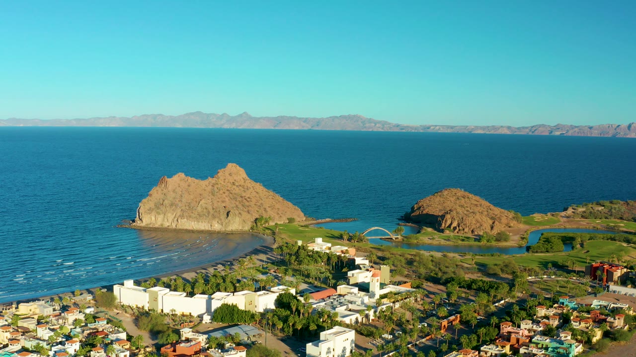 Mid-clip aerial frame over Loreto showing the rocky outcrops slightly lower in the composition, a broad band of calm ocean, white town blocks, green land near the water, and no visible people.