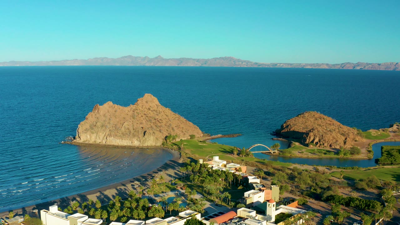 Opening aerial frame over Loreto, Baja California Sur showing blue ocean, a sandy beach, tan rock formations, white buildings, palms, distant mountains, and no visible people.