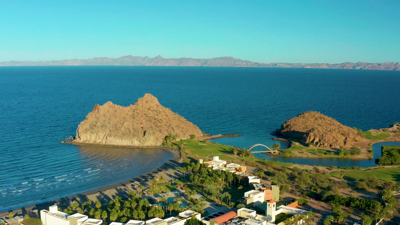 Aerial view in Loreto, Baja California Sur with the main tan rock centered, the curved beach at lower left, a small bridge near green water, distant mountains, and no visible people.