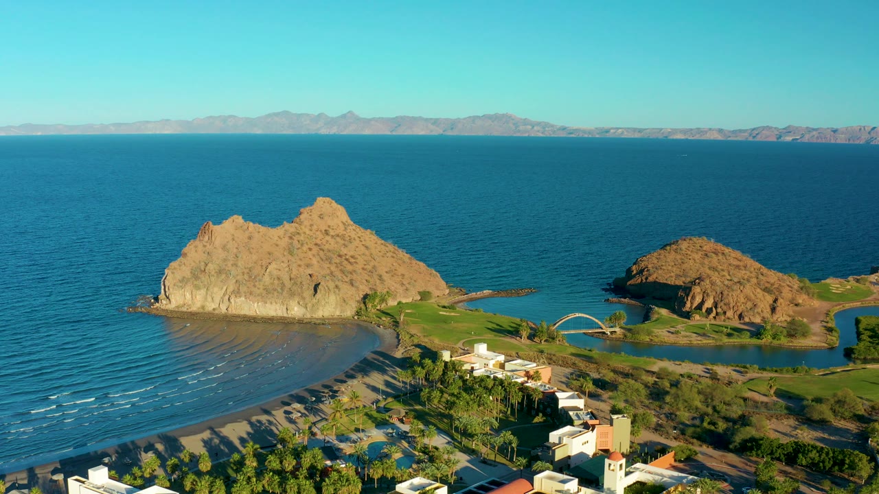 Aerial beach scene with tan rock, ocean, bridge, greenery, and buildings.