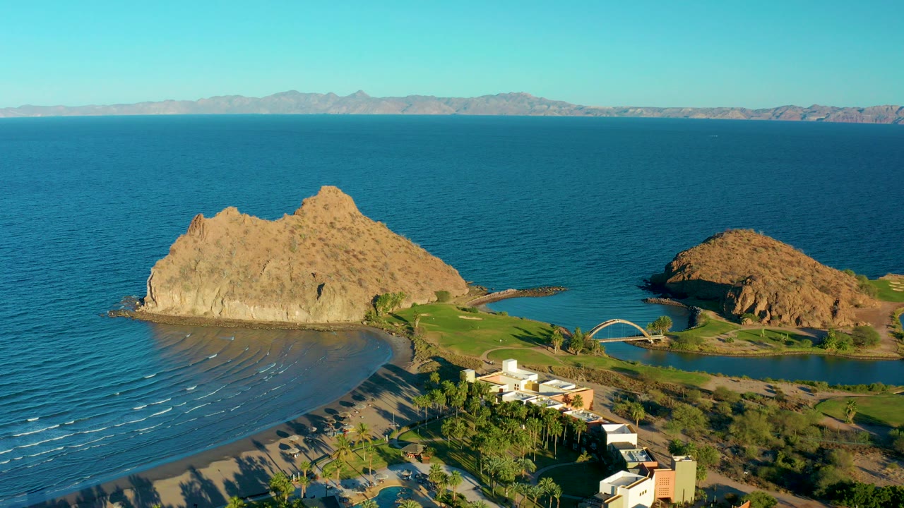 Aerial frame from Loreto, Baja California Sur showing tan rocky outcrops beside calm blue ocean, a crescent beach, resort buildings, green landscaping, distant mountains, and no visible people.