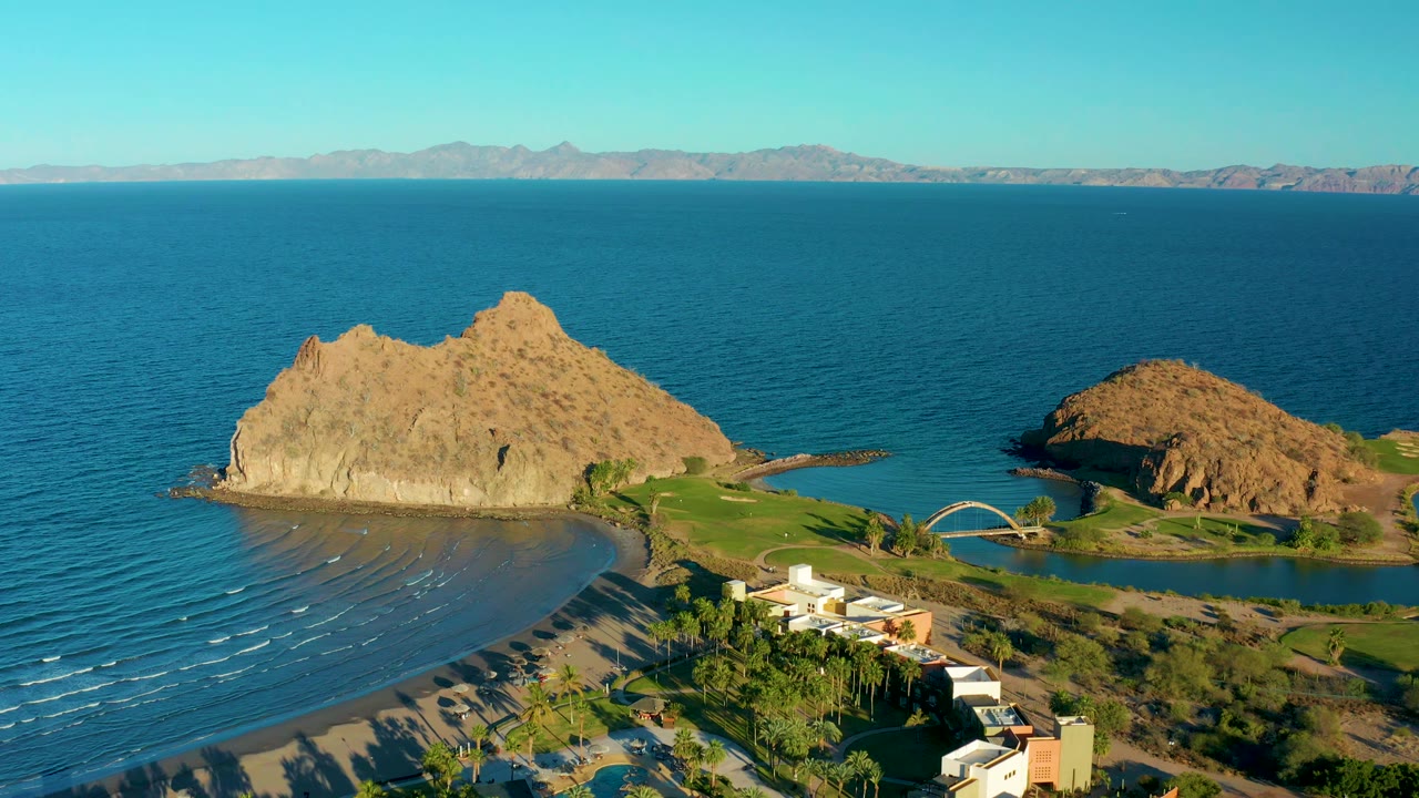 A second aerial frame over Loreto, Baja California Sur keeps the sandy shoreline and resort roofs in the lower frame while tan rock formations sit against calm ocean and no visible people.