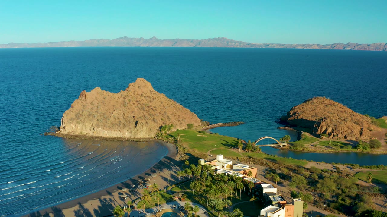 Aerial view in Loreto, Baja California Sur with the main rocky outcrop centered, smaller rock to the right, resort landscaping below, visible wave lines near shore, and no visible people.