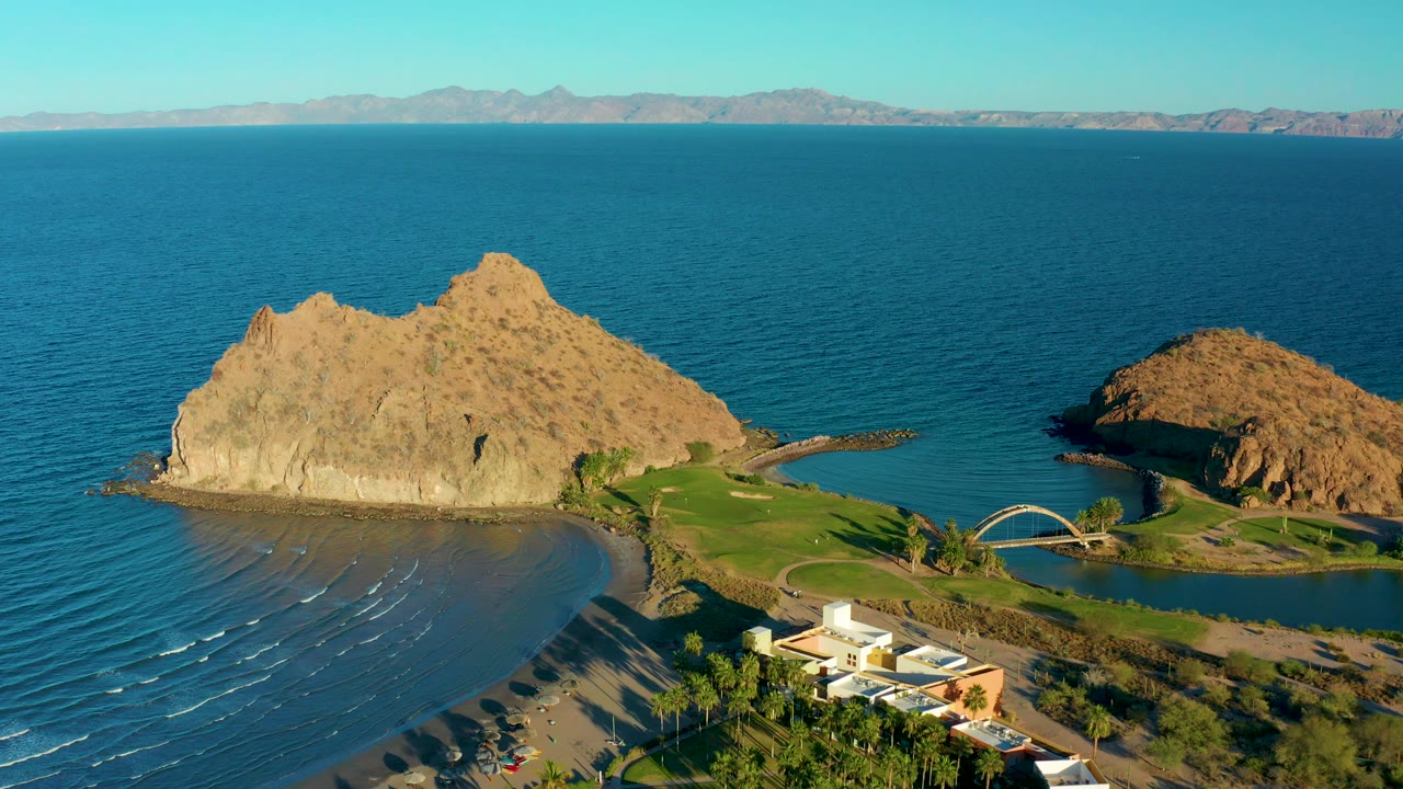 Rocky island and beach seen from above with waves and a small bridge.