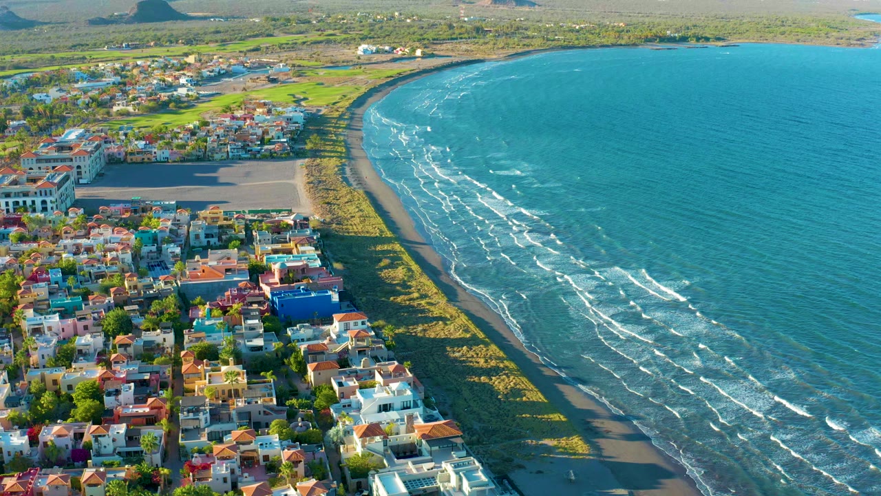 At 0:00, an elevated view over Loreto, Baja California Sur, Mexico shows colorful coastal homes, a curved sandy beach, turquoise water, distant low hills, and no visible people.