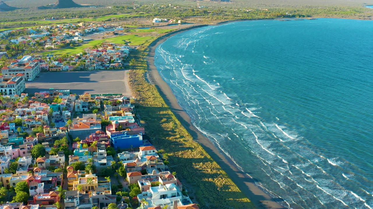 Aerial view over Loreto with the town shifted slightly along the left edge, a pale beach curve below, wind-lined blue water to the right, and no visible people.