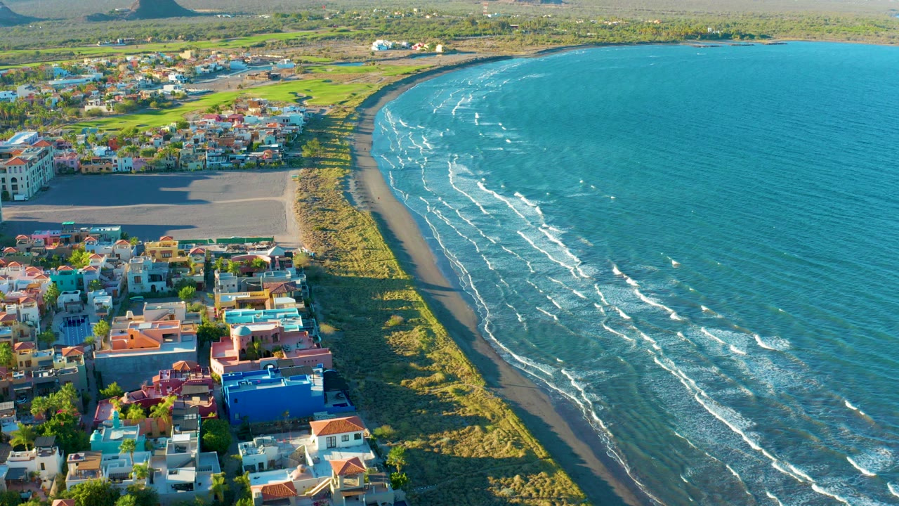 Aerial frame over Loreto, Baja California Sur, showing colorful homes near a sandy curved beach, turquoise water with thin white wave lines, green inland areas, distant hills, and no visible people.