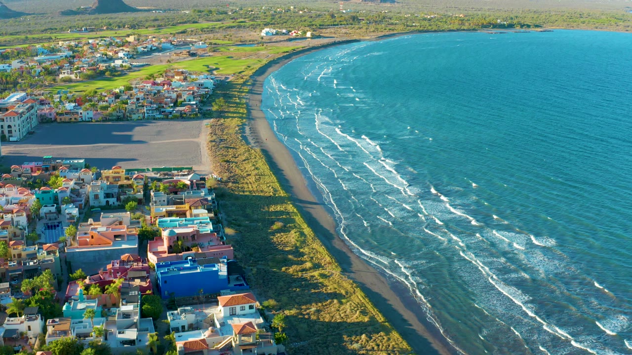 Drone view of beachside houses, green shoreline, and blue water with small waves.