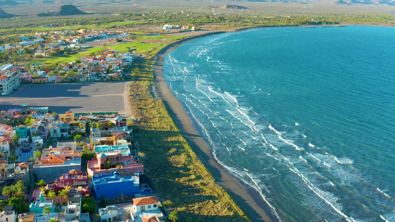 High aerial frame of Loreto showing more open water to the right, the town and rooftops along the lower left, a curved sand beach, green fields beyond the shoreline, distant hills, and no visible people.