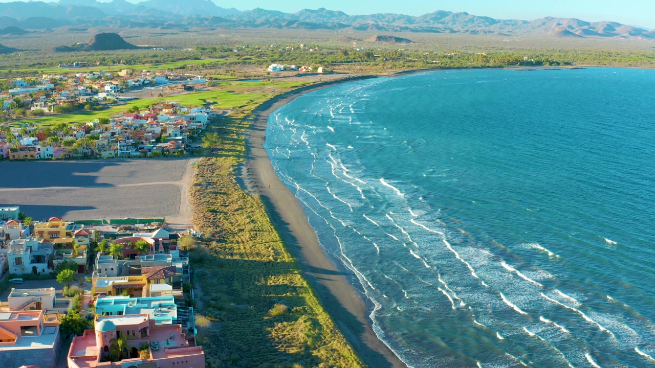 Opening aerial frame over Loreto, Baja California Sur, with colorful homes at lower left, a narrow sandy beach curving beside turquoise surf, distant mountains on the horizon, and no visible people.