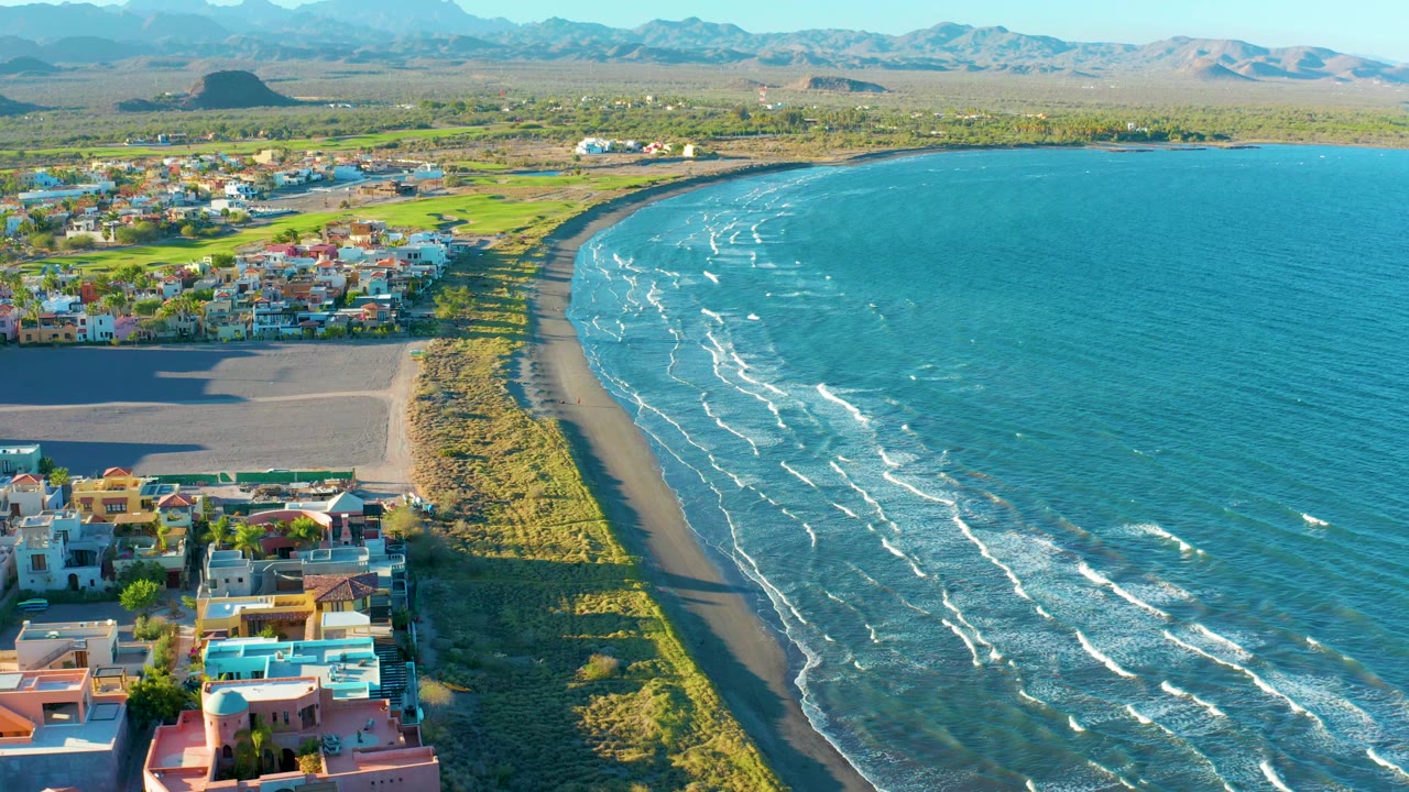 Aerial coastline view with beach homes, surf lines, grassy dunes, and mountains.