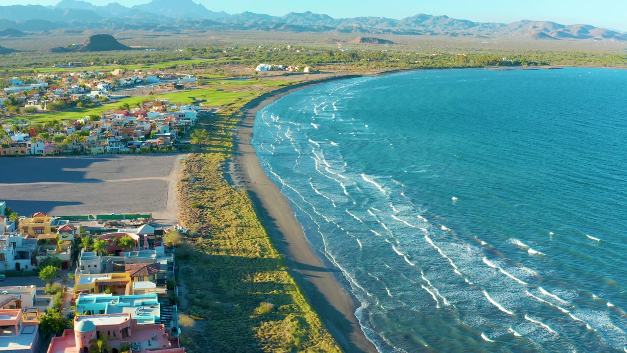 Loreto aerial frame with the sandy beach and grassy dune strip forming a long curve, bright blue ocean filling the right side, clustered town buildings to the left, distant mountains, and no visible people.